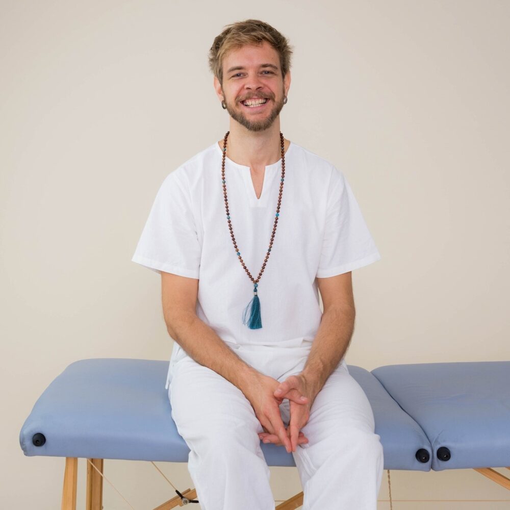 Friendly looking medical massage therapist sitting on light blue massage table smiling towards the camera, wearing white clothes and a mala.