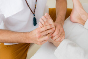 Guy in white shirt and prayer mala massaging another guys foot performing a foot reflexology.