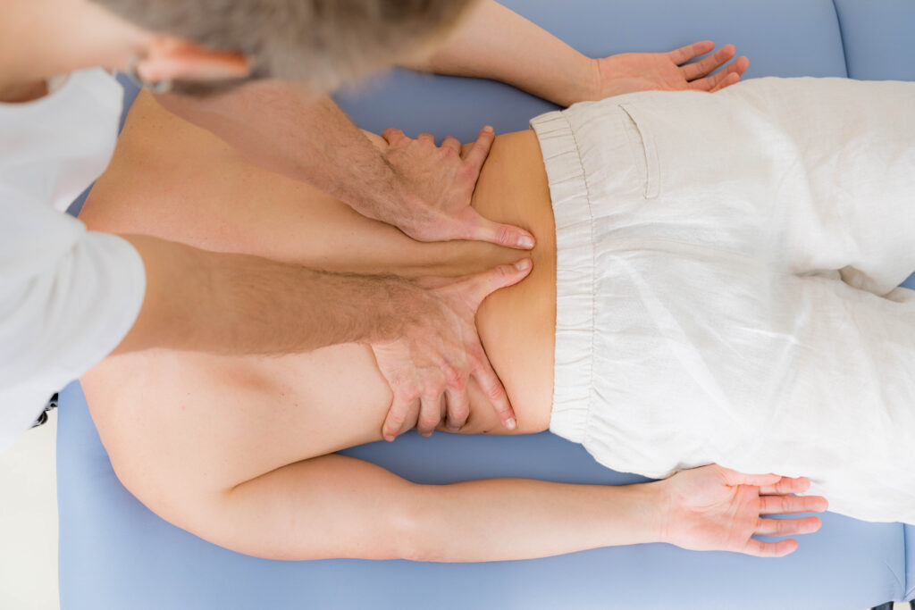 A guy laying on a light blue massage table while the massage therapist massages the back with oil and both hands parallel on the lower back.