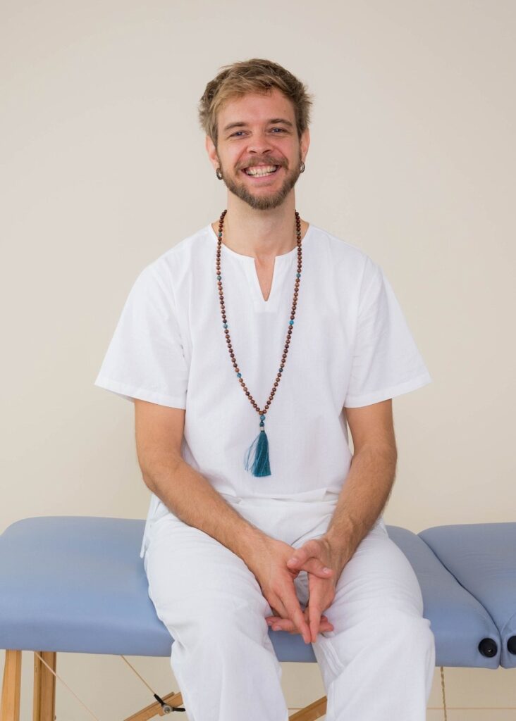 Guy sitting on a light blue massage table wearing white clothes and a mala, smiling to the camera.