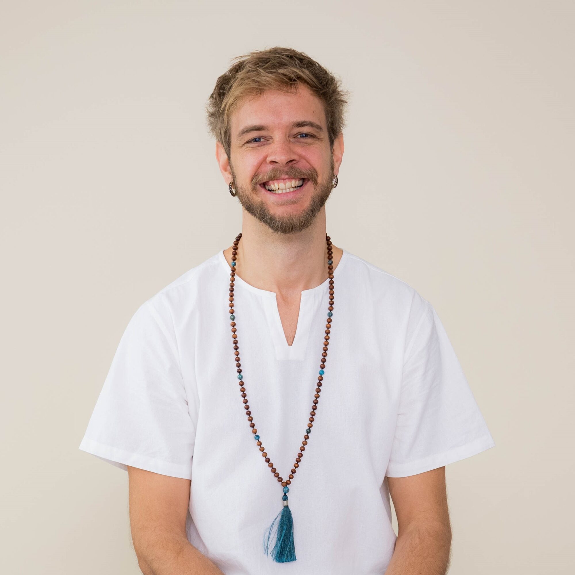 Guy sitting on a light blue massage table wearing white clothes and a mala, smiling to the camera.