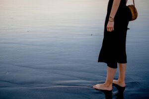 Woman in black dress wearing a bamboo purse standing barefooted on the beach with black sand.