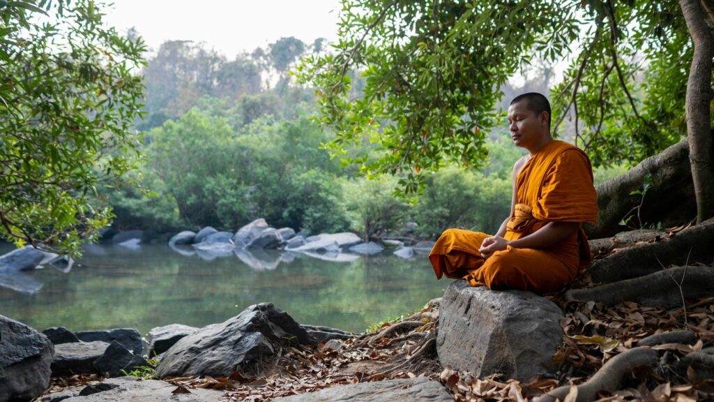 Monk in orange robes meditating in Thailand next to a river and big trees.